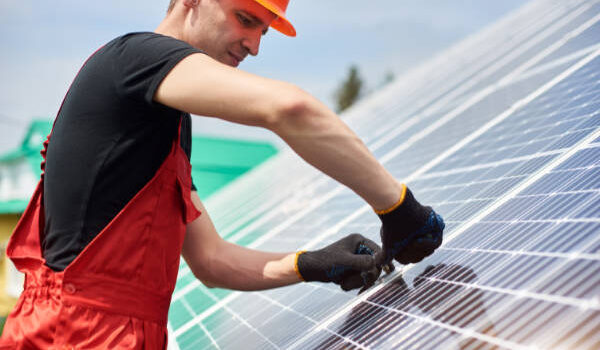 Electrician installing solar panels to save energy. A man is wearing an orange uniform, hard hat, black t-shirt and gloves against the blue sky. Alternative energy sustainable concept